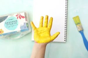 Hand with bright yellow paint on palm and fingers resting above a white notebook, with a paintbrush and art supplies nearby on a light blue surface for this handprint art
