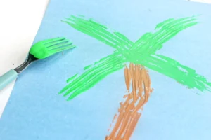 Blue paper with bright green cross-like brushstrokes and a brown vertical section to look like a palm tree, a green-tipped fork resting at the left edge.