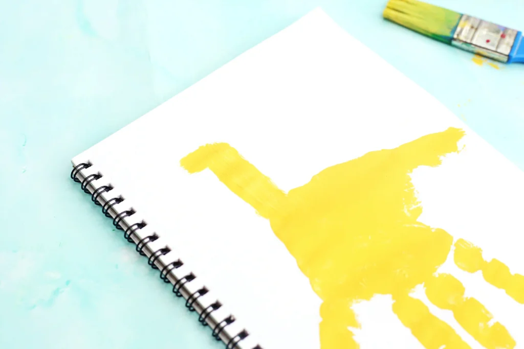Notebook with spiral binding on a pale blue surface; white page has yellow handprint paint smear and a paint tube in the corner (art setup) for this G is for Giraffe Handprint craft