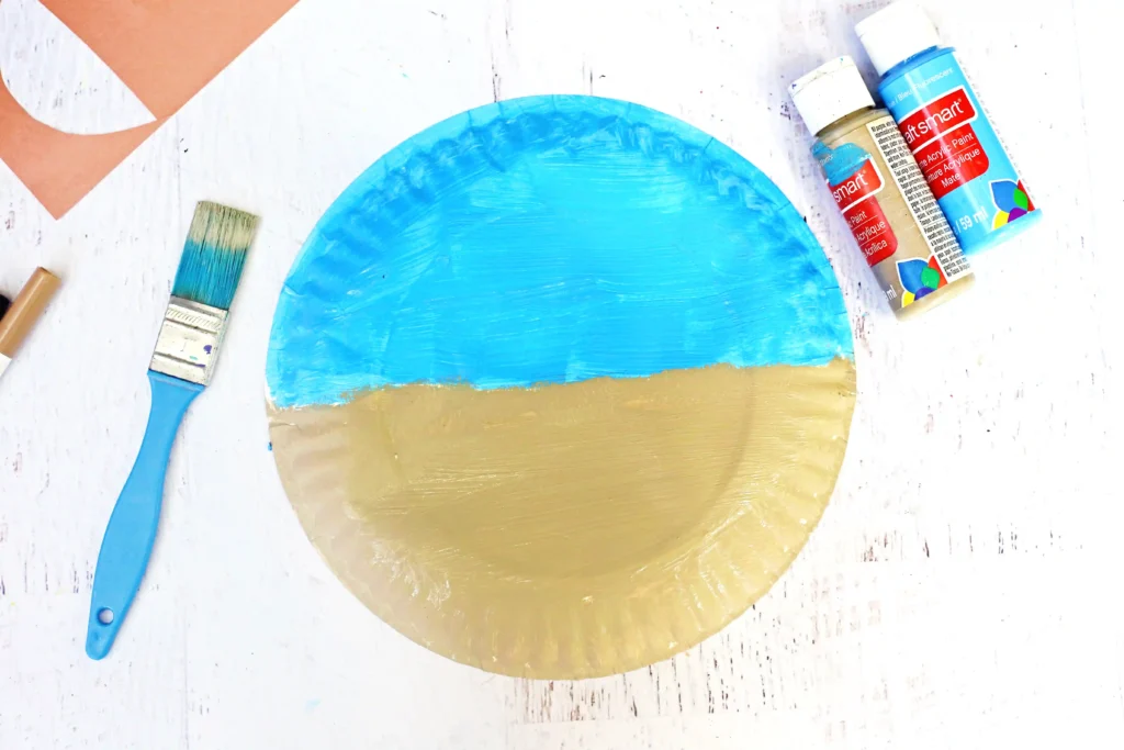 Artist's workspace with a blue and tan-painted paper plate, two brushes, and acrylic paint tubes on a white wooden surface.
