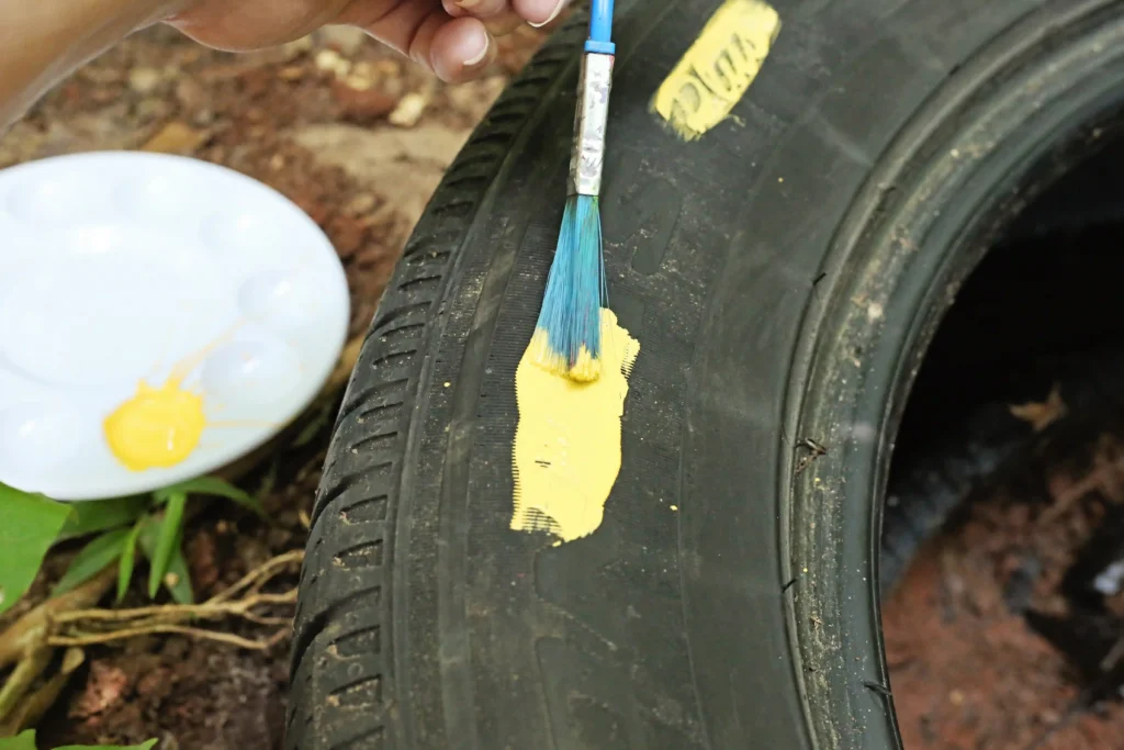 Person applying yellow paint to the tire sidewall with a brush; a paint plate with yellow paint sits nearby.