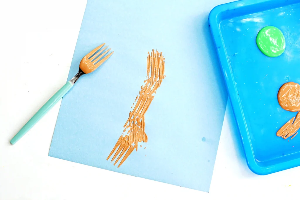 Light blue paper with brown paint scraped by a fork, plus a turquoise-handled brush and a blue paint tray with a green paint blob on the right.