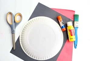 supplies for the paper plate craft: scissors, white paper plate, colored papers, glue sticks, and a paintbrush on a pink and gray backdrop.