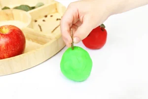 Hand picking a green playdough apple from a display of red and green apples near a wooden basket.