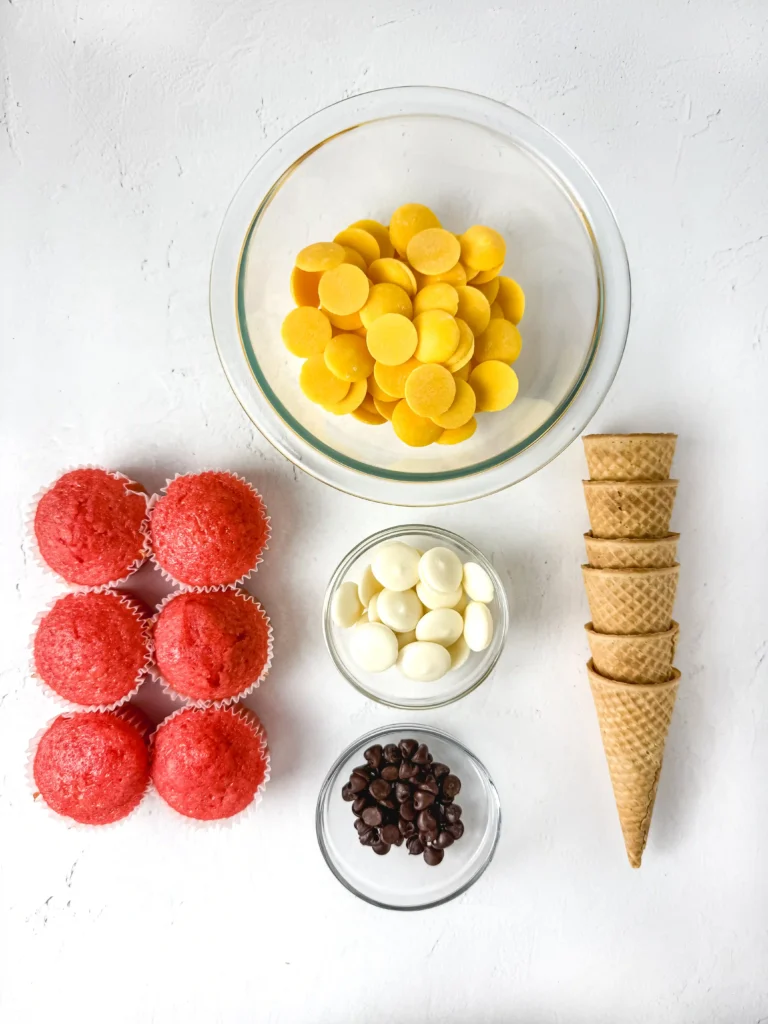 ingredients for this back to school dessert- Top-down view of assorted sweets: yellow candy discs in a glass bowl, red cupcakes, white chocolate chips, stacked waffle cones, and chocolate chips on a white surface.