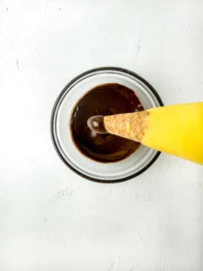 Overhead shot of a white plate with dark chocolate and a yellow dipped ice cream cone partially submerged in the chocolate, on a pale background.