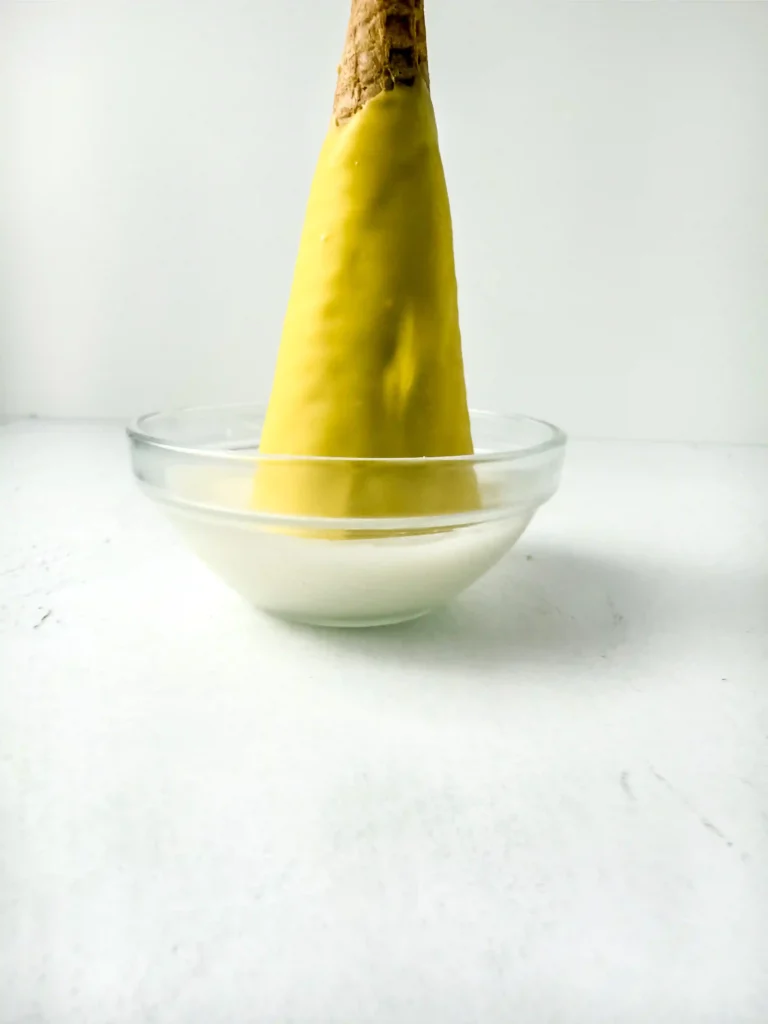 Ice cream cone dipped in yellow melted chocolate being dipped into a bowl with white melting chocolate for these teacher appreciation gifts.