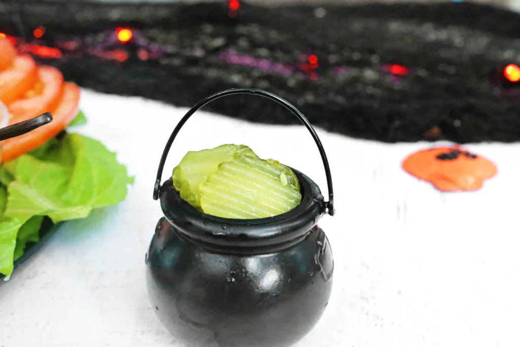 Close-up of a black cauldron with a curved metal hoop around the neck, holding a pickle slice inside; blurred greens on the left and an orange garnish on the right.