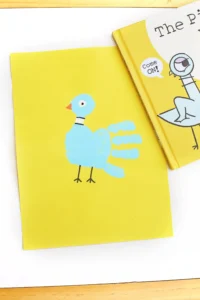 a pigeon handprint craft that is a blue handprint on yellow paper with details to look like a pigeon next to the book "The pigeon has to go to school"