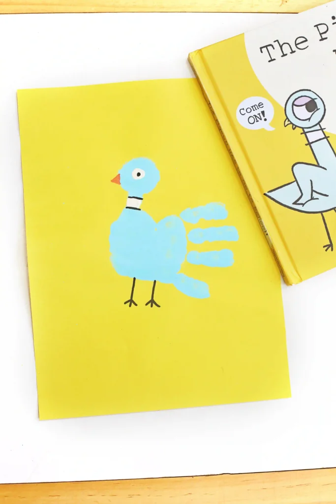 a pigeon handprint craft that is a blue handprint on yellow paper with details to look like a pigeon next to the book "The pigeon has to go to school"