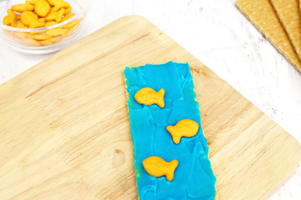 Ocean Graham Crackers - Cultivating Brilliant Minds Blue icing on a graham cracker on a wooden board topped with goldfish crackers