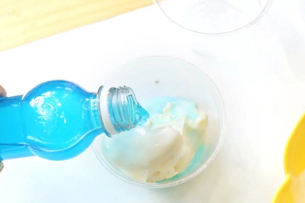 Blue liquid pours from a blue bottle into a clear cup on a white surface, with a hint of ice inside the cup.