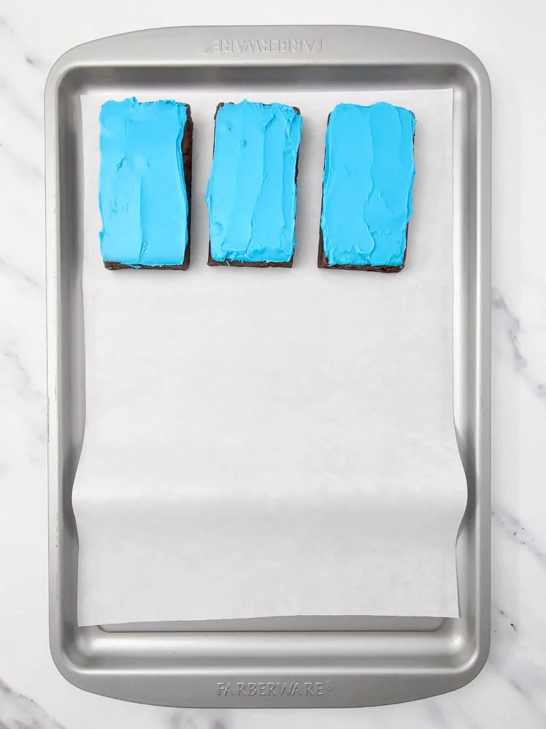 Three rectangular brownies with bright blue frosting arranged in a row on parchment in a metal baking tray