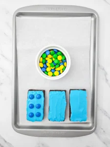 Top-down view of a metal baking tray with three blue-frosted brownies with blue M&Ms and a white bowl of colorful candy-coated chocolates.