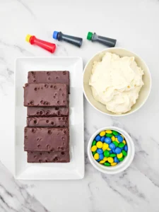 ingredients for the lego brownies- Chocolate Cosmic brownies with the sprinkles removed on a white plate beside a bowl of creamy frosting and a small dish of colorful M&M candies on a marble surface.