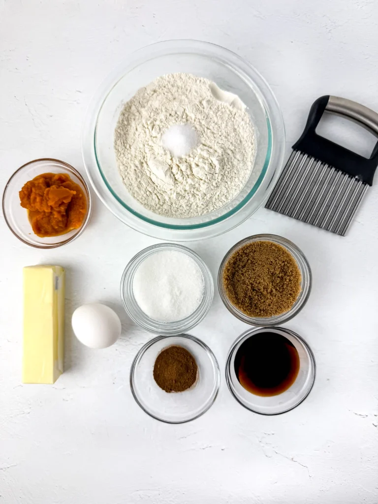 ingredients for the pumpkin spice cookie fries: a glass bowl of flour with a small mound of salt in the center, surrounded by butter, brown sugar, white sugar, an egg, vanilla, pumpkin puree, and a pastry brush.
