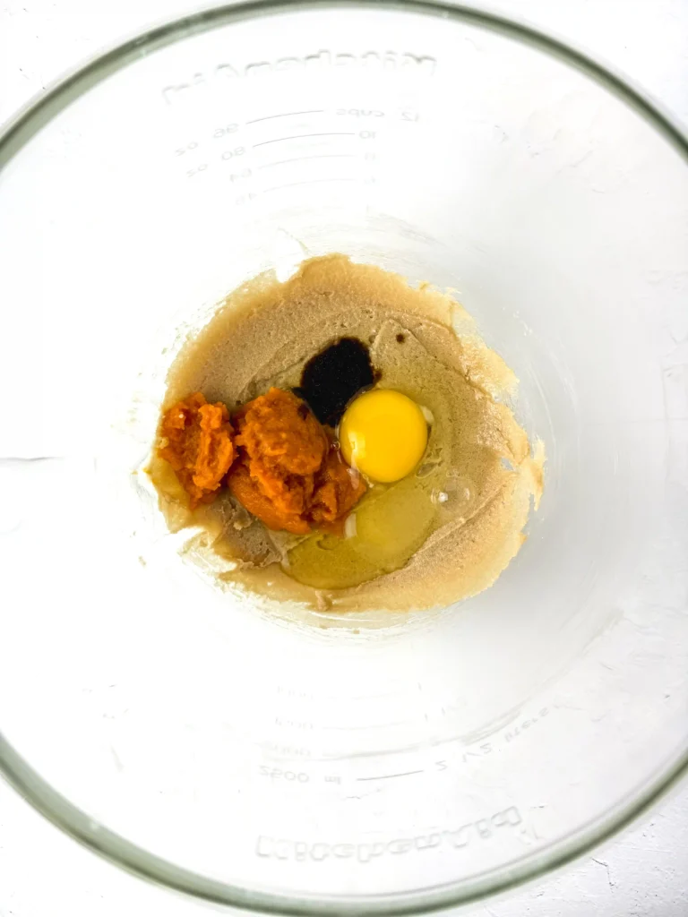 Top-down view of a glass bowl with baking dough and ingredients: an egg, brown sugar, and orange pumpkin puree ready to mix for these pumpkin cookies