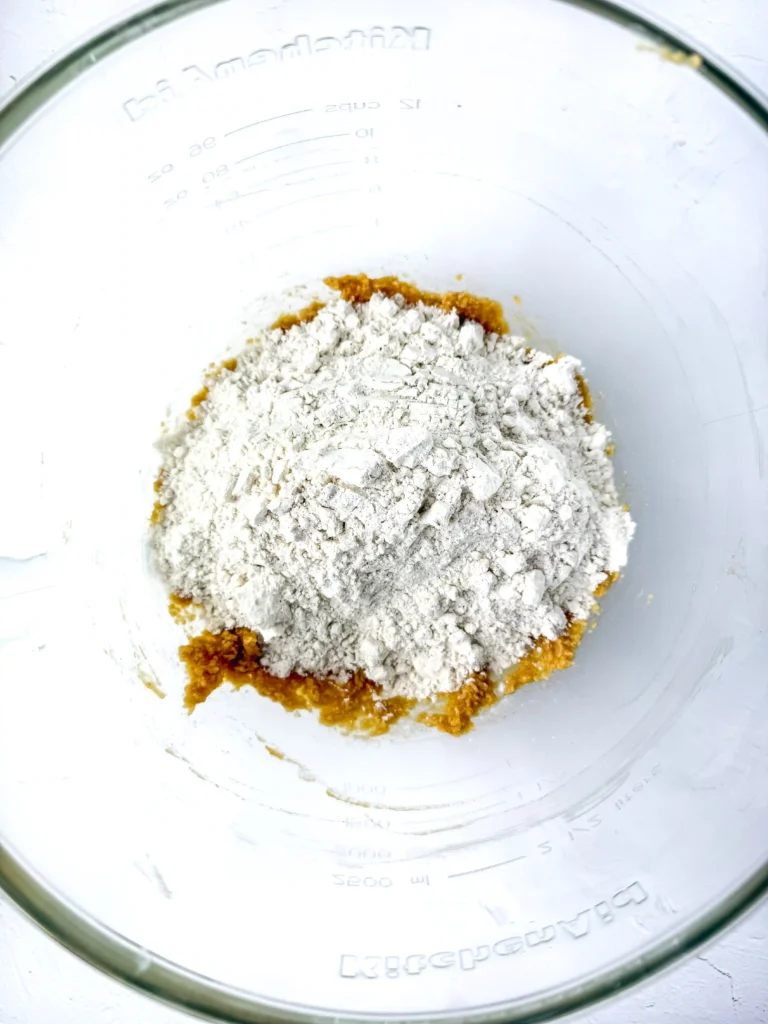 Clear glass bowl containing orange batter or dough, topped with a mound of white flour.