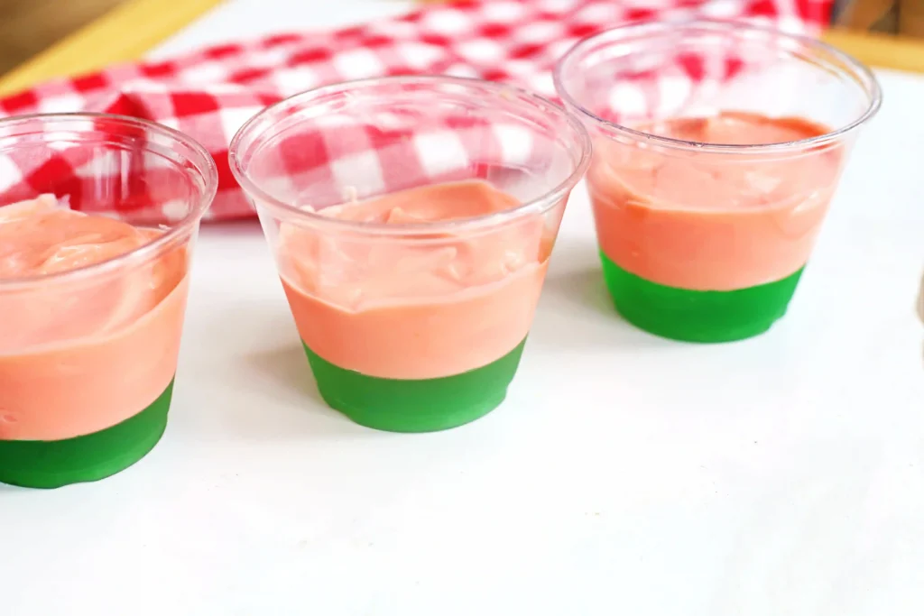 Three clear cups hold pink vanilla pudding layered with a bright green jello bottom on a white surface.