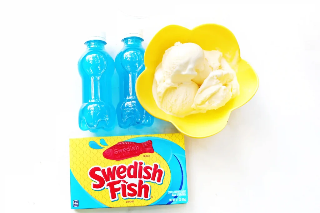 ingredients for this easy summer treat- Two blue beverage bottles beside a bright yellow bowl filled with vanilla ice cream, with a Swedish Fish candy box at the bottom edge.