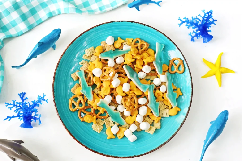 Turquoise plate of ocean-themed snacks, including blue shark-shaped candies, pretzels, cereal squares, and marshmallows on a white table with blue decor nearby. (informative)