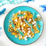Turquoise plate of ocean-themed snacks, including blue shark-shaped candies, pretzels, cereal squares, and marshmallows on a white table with blue decor nearby. (informative)