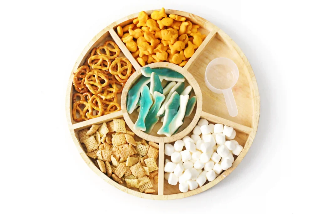 a tray with pretzels, goldfish, gummy sharks, chex mix, and mini marshmallows for a shark trail mix