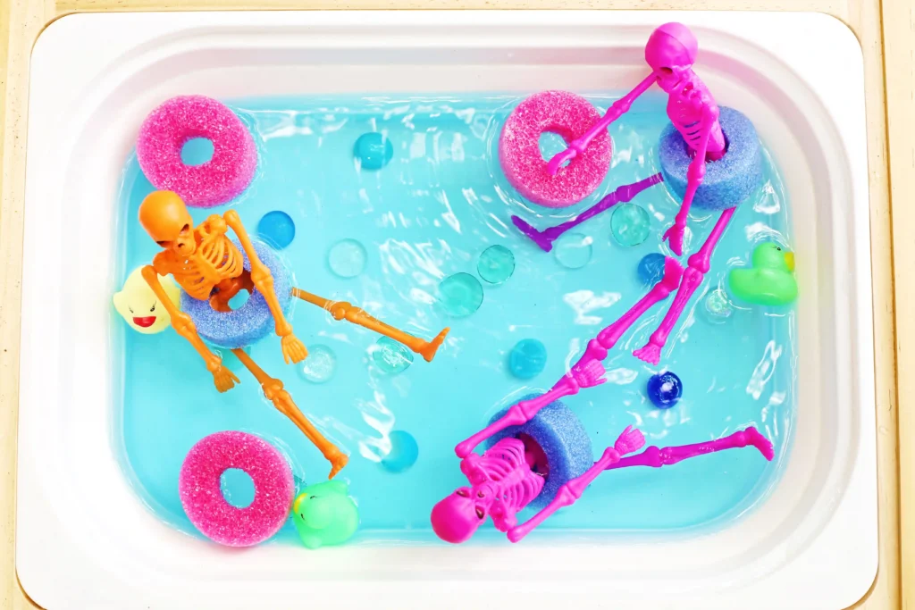 a skeleton pool party sensory bin that is a bin filled blue water that has pool noodle slices, rubber ducks, and plastic skeletons