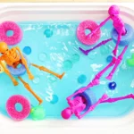 a skeleton pool party sensory bin that is a bin filled blue water that has pool noodle slices, rubber ducks, and plastic skeletons