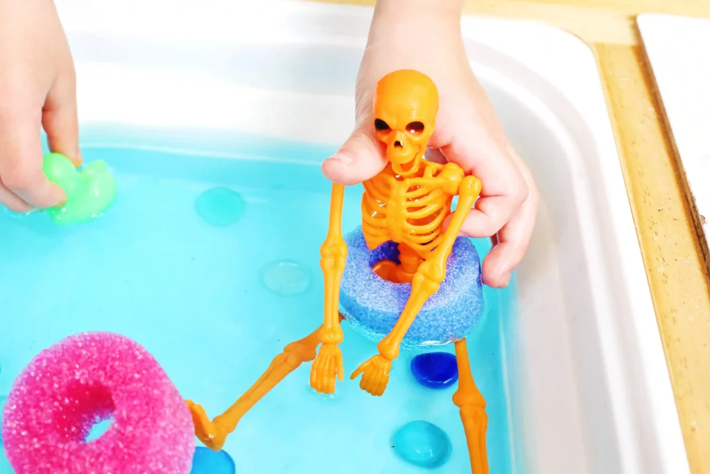 Orange plastic skeleton being held over a blue toy pool with colorful foam end toys nearby (green duck, pink pool noodle slice).