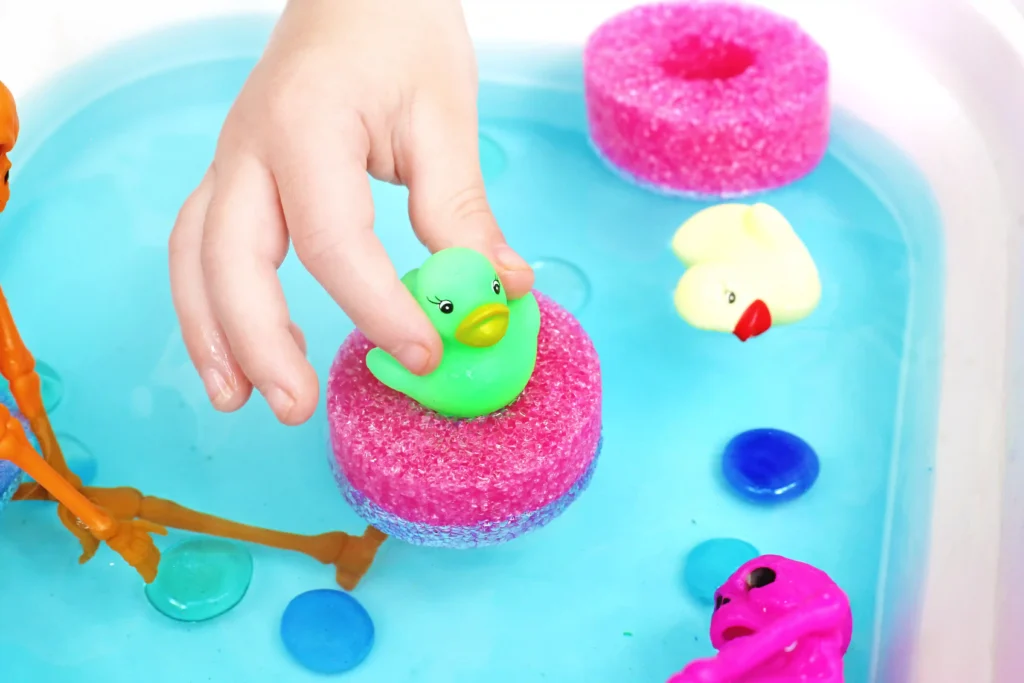 Child's hand placing a green rubber duck onto a pink foam float in blue bath water.