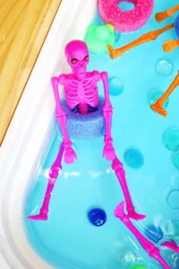 Pink plastic skeleton toy partially submerged in a bright blue pool noodle slice, with other colorful pool toys nearby