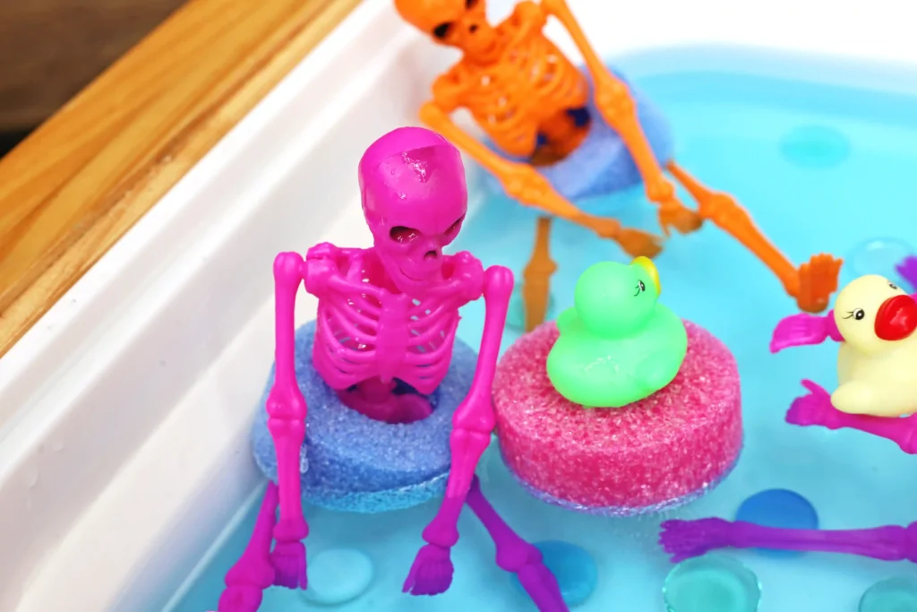 Pink plastic skeleton toy in a blue bath with colorful pool noodle slices and rubber ducks nearby.