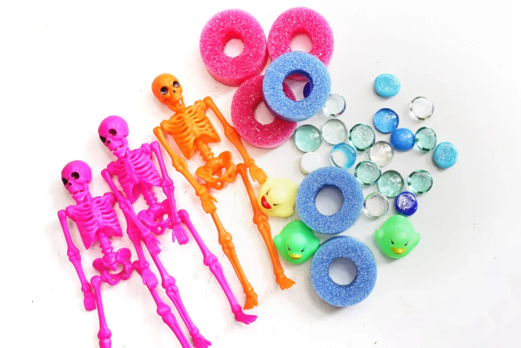 supplies for this summerween activity - Assorted colorful plastic skeletons, toy ghosts, foam donuts, and glass pebbles arranged for a playful Halloween-themed display.