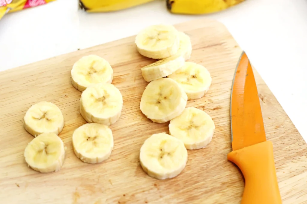 Banana slices arranged on a wooden cutting board with an orange-handled knife nearby.