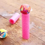 Pink foam pool noodle upright with a colorful patterned ball balanced on top, on a weathered wooden deck.