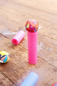 Pink foam pool noodle upright with a colorful patterned ball balanced on top, on a weathered wooden deck.