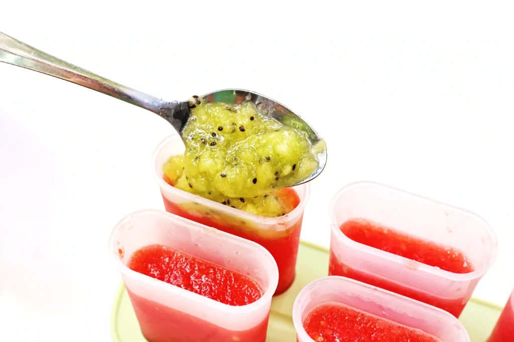 Watermelon Kiwi Popsicles - Cultivating Brilliant Minds Spoon scooping bright green kiwi topping into popsicle molds with blended watermelon on a light tray in the background.