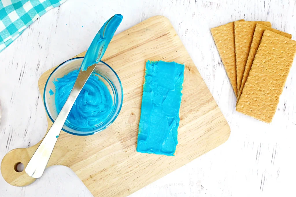 Ocean Graham Crackers - Cultivating Brilliant Minds Blue frosting being spread from a knife onto a graham cracker on a wooden cutting board, with crackers nearby in the background for these under the sea graham crackers