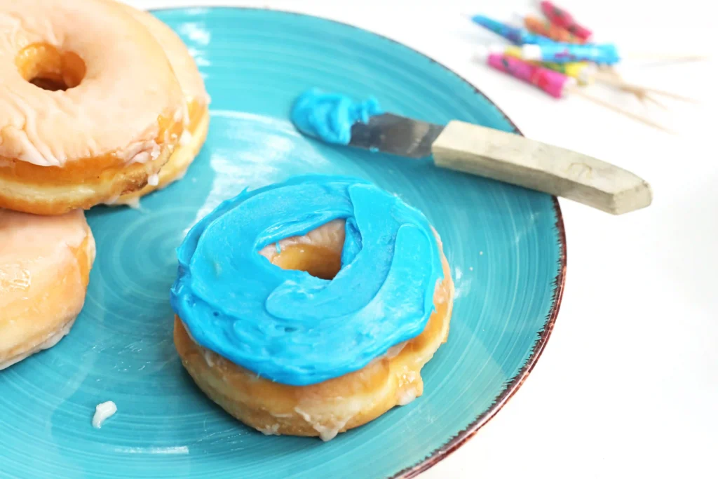 Donuts with bright blue frosting on a turquoise plate, with a knife and piping bag nearby and colorful clothespins in the background.
