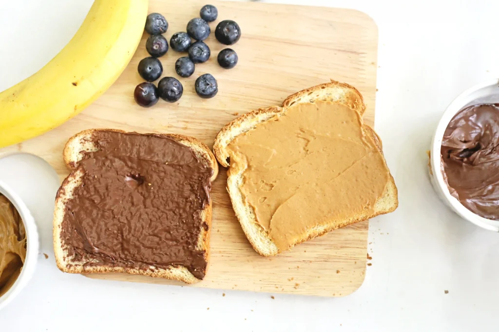 bread with peanut butter and chocolate hazelnut spread on a wooden cutting board next to blueberries and a banana