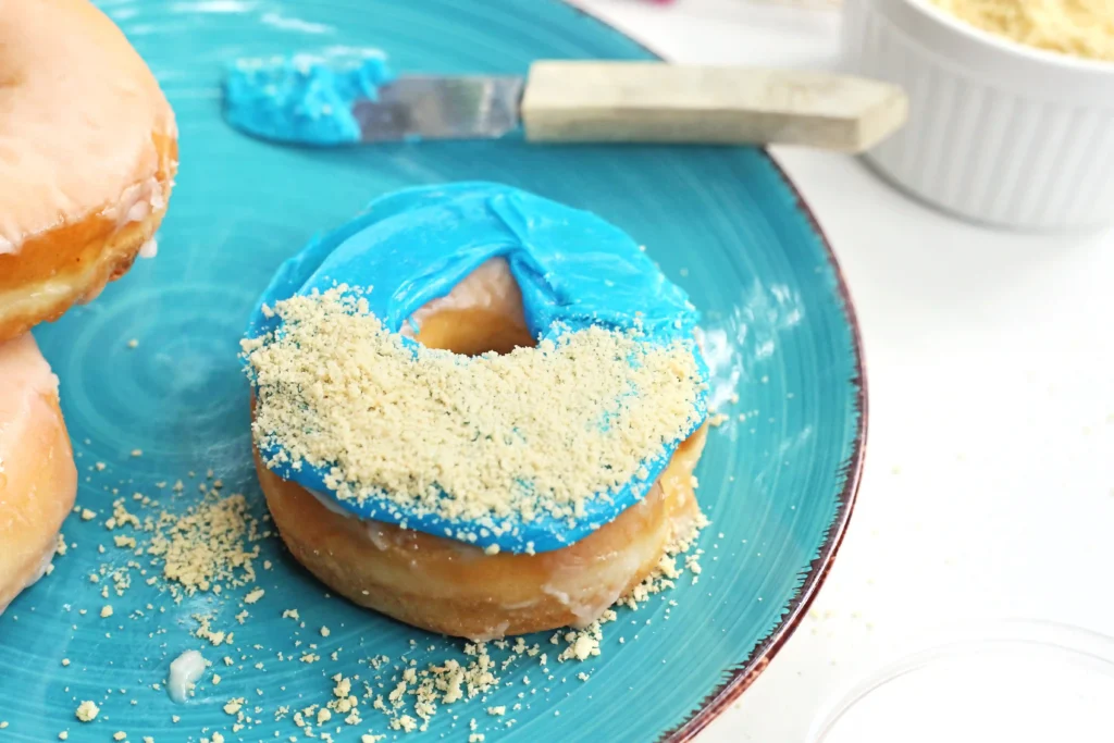 Blue-frosted donut with crumb topping on a blue plate, with a spatula in the background.