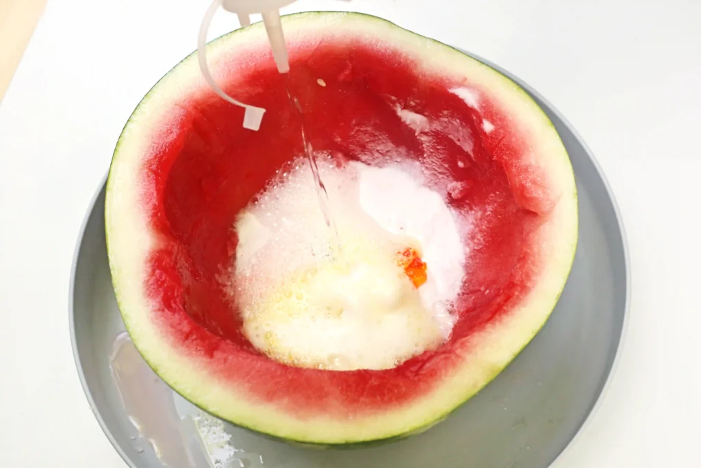 a person dripping vinegar from a squeeze bottle into a hollowed out watermelon with baking soda and food coloring