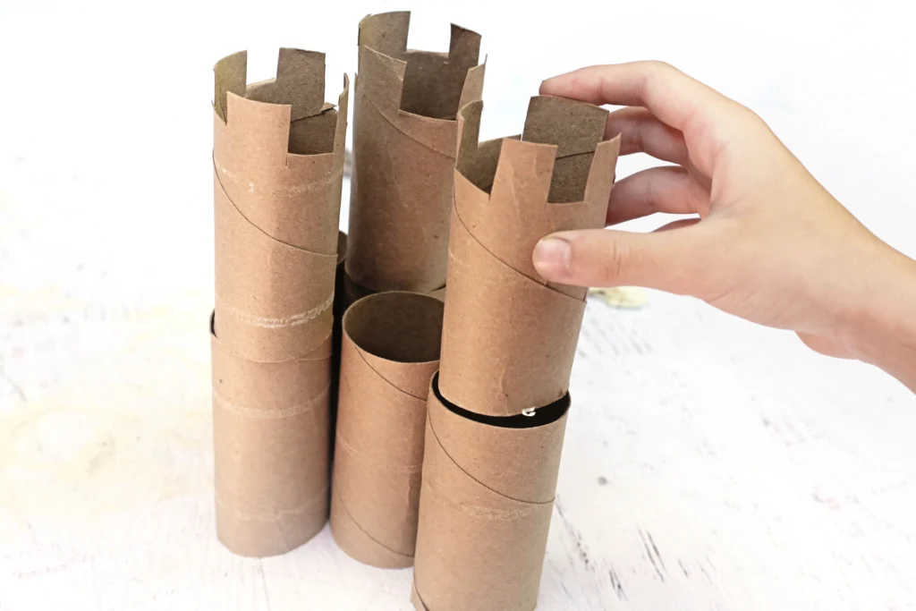 Hand placing cardboard toilet-paper tubes arranged as a small castle, on a light wooden surface.