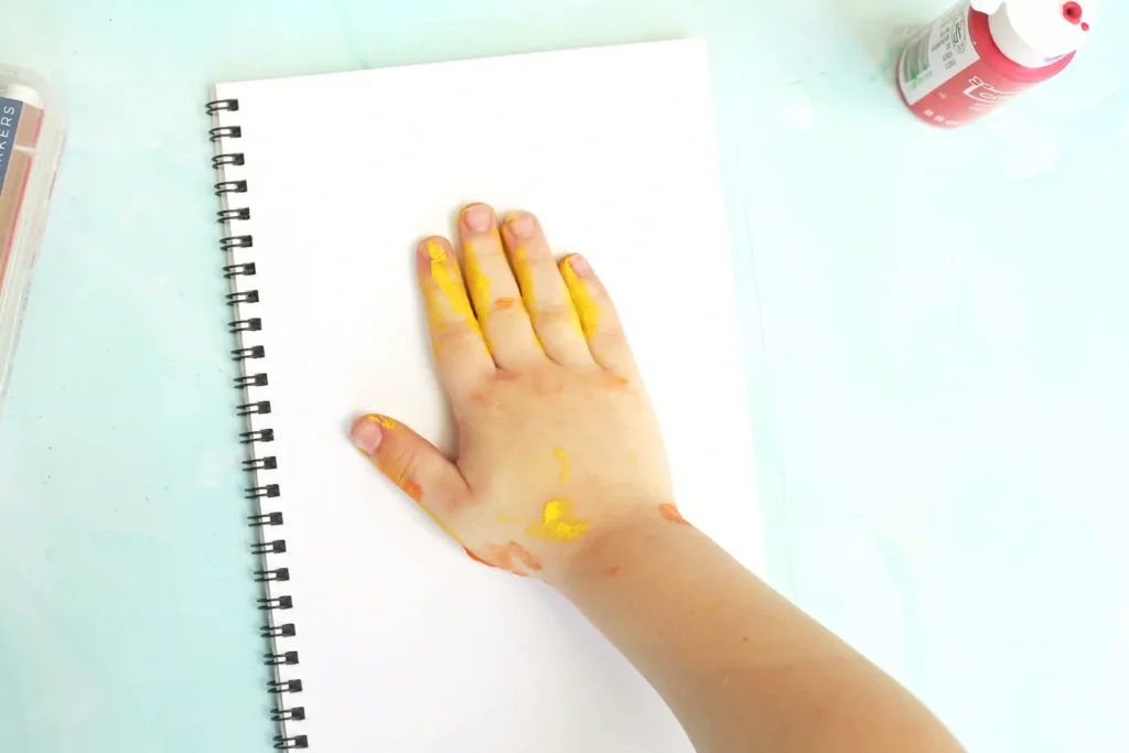 Child's hand covered in yellow paint resting on a blank notebook page with a spiral binding and a paint bottle nearby on a light tabletop for this campfire handprint craft
