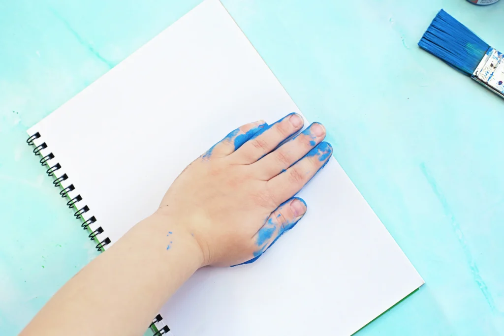 Hand with blue paint on fingers rests on a white spiral notebook on a turquoise painting surface, with a blue paintbrush to the right.