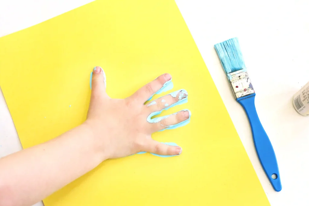 Hand smeared with blue paint resting on a bright yellow sheet, with a blue paintbrush nearby for these mo willems activities for preschool