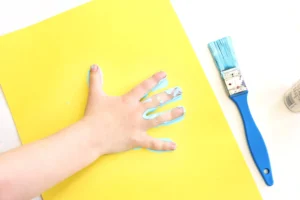 Hand smeared with blue paint resting on a bright yellow sheet, with a blue paintbrush nearby for this mo willems activity for preschool