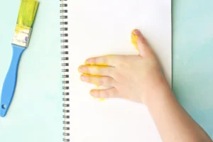 Hand smeared with yellow paint presses down on a blank notebook page next to a blue-handled paintbrush on a pale green surface for this handprint craft for kids