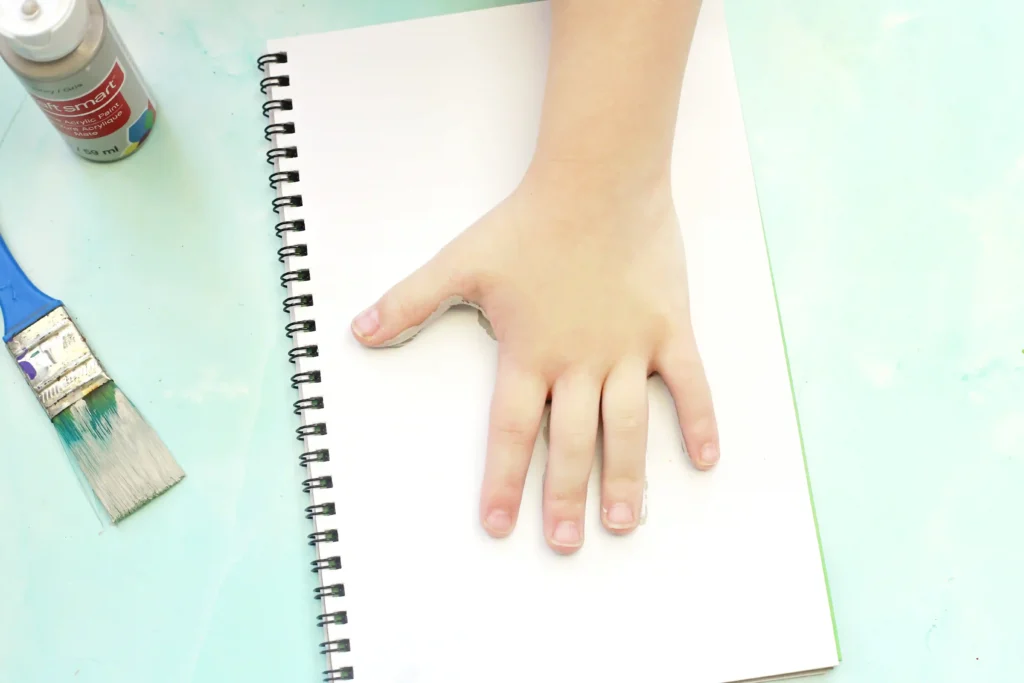 Letter E Elephant Handprint Craft - Cultivating Brilliant Minds Small hand resting on a blank spiral notebook page with a paintbrush and acrylic bottle nearby on a pale blue surface for this elephant handprint craft